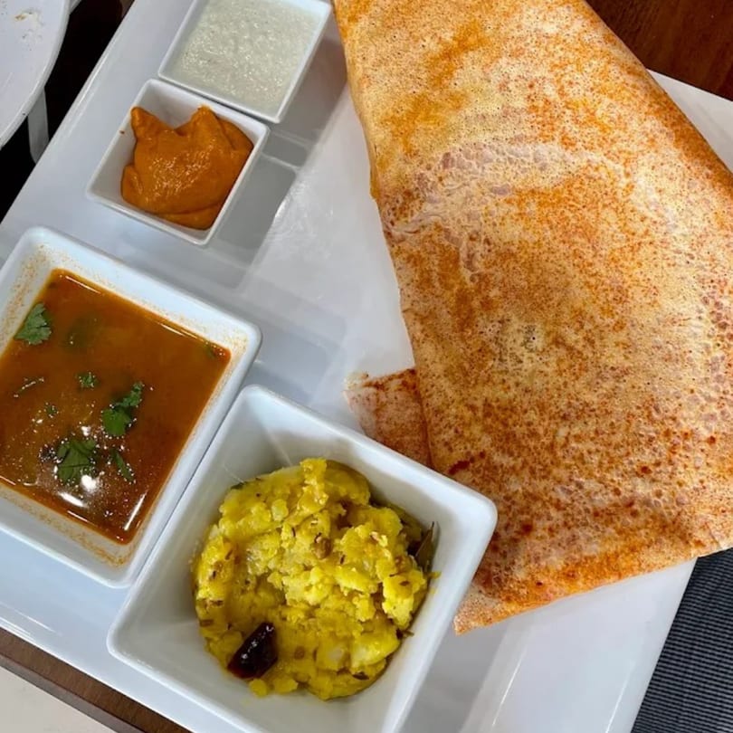 A golden-brown dosa with four small white bowls of chutneys and sambar on a white plate.
