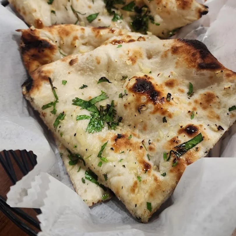 Close-up of golden-brown garlic naan bread, garnished with fresh green herbs, served in a basket.